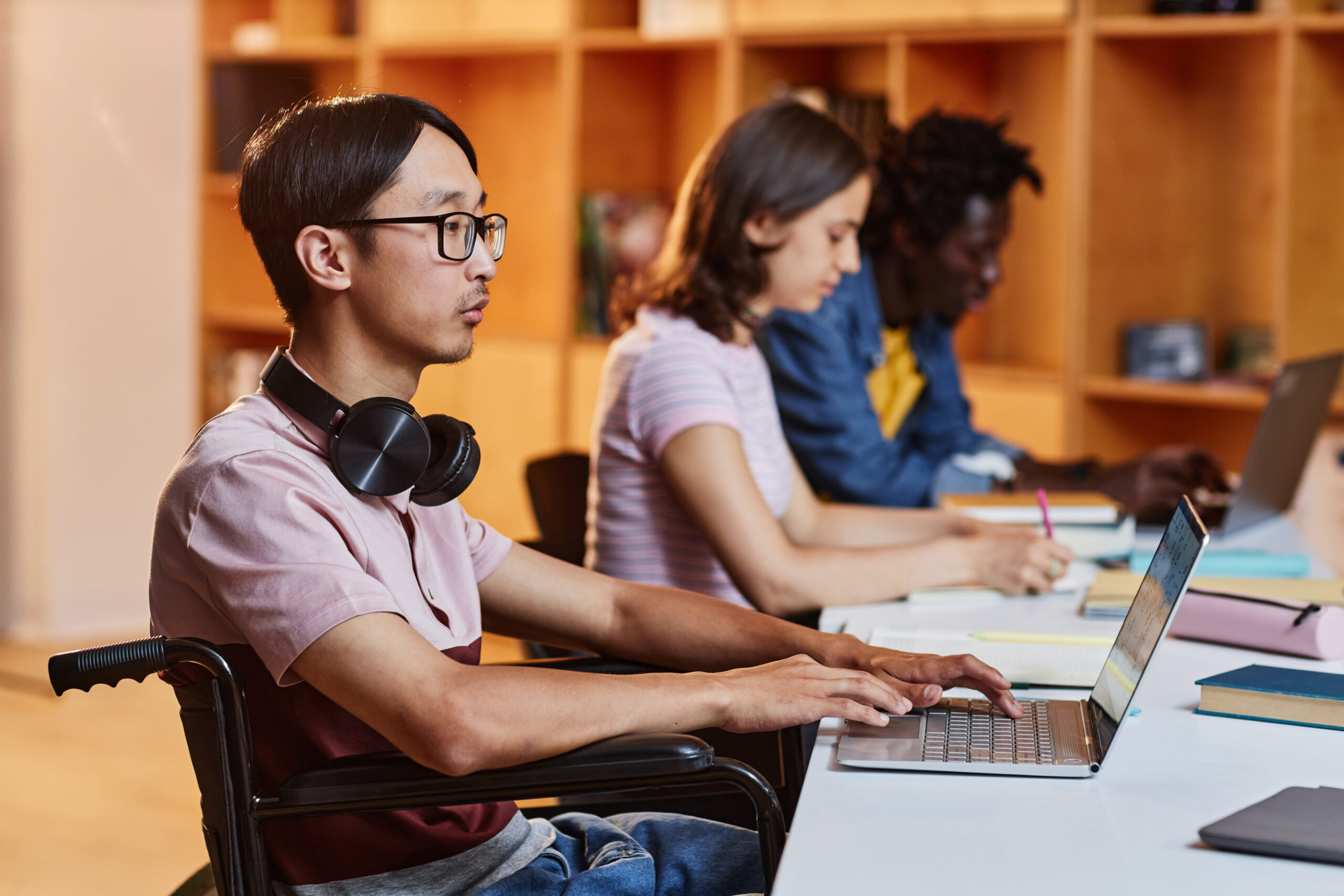 comunicação inclusiva representada por pessoas de diferentes etnias. em primeiro plano, um jovem asiático cadeirante mexe em seu notebook. ao fundo, uma mulher jovem, branca, e mais ao fundo, um homem também jovem, negro